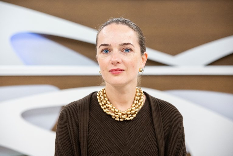 Mulher de olhos azuis intensos, cabelo castanho claro preso, vestindo um suéter marrom e um colar dourado volumoso, com um fundo desfocado de formas abstratas brancas. Seu olhar transmite seriedade e profundidade, características que podem ser relacionadas à discussão sobre o futuro da democracia brasileira.